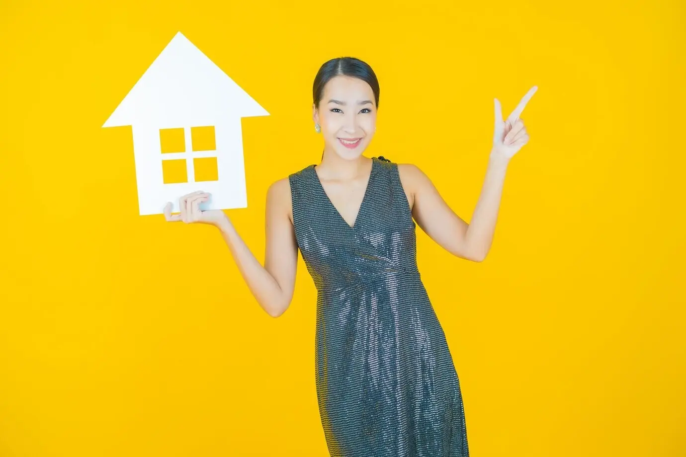 Портрет красивой молодой азиатской женщины с бумажным символом дома на жёлтом фоне.